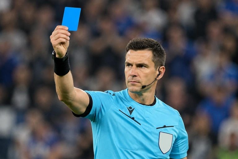 Arbitre de football professionnel levant un carton bleu lors d'un match, portant un maillot officiel et un casque micro.